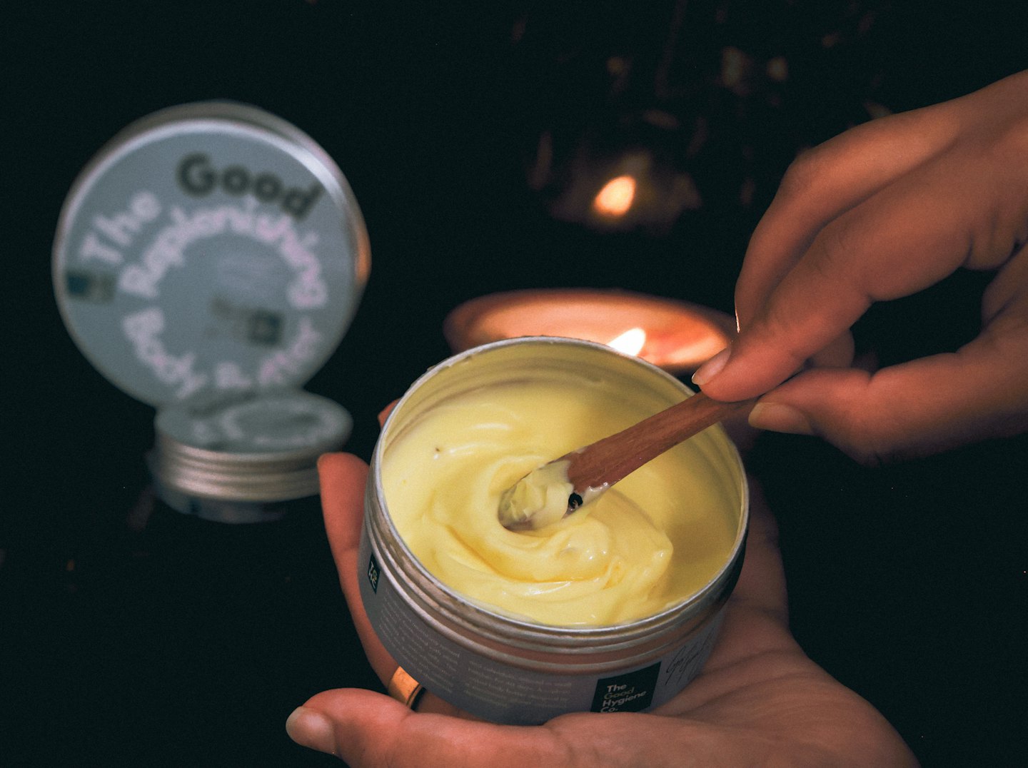 Hands use a wooden spatula to scoop yellow body butter.